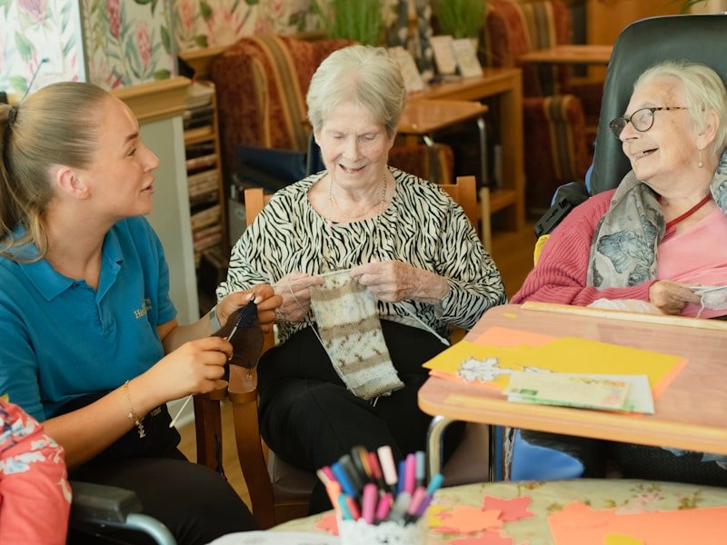 A senior knitting at a sunny table with a caregiver assisting